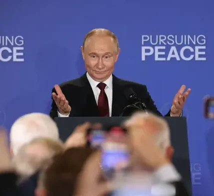 Путін за трибуною з піднятими руками на тлі напису Pursuing Peace