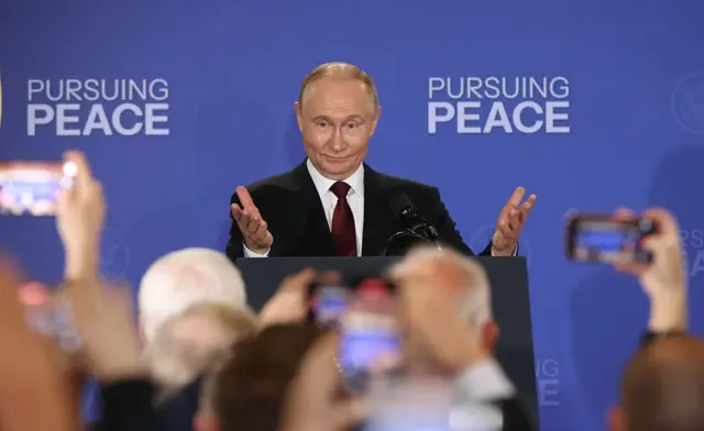 Путін за трибуною з піднятими руками на тлі напису Pursuing Peace