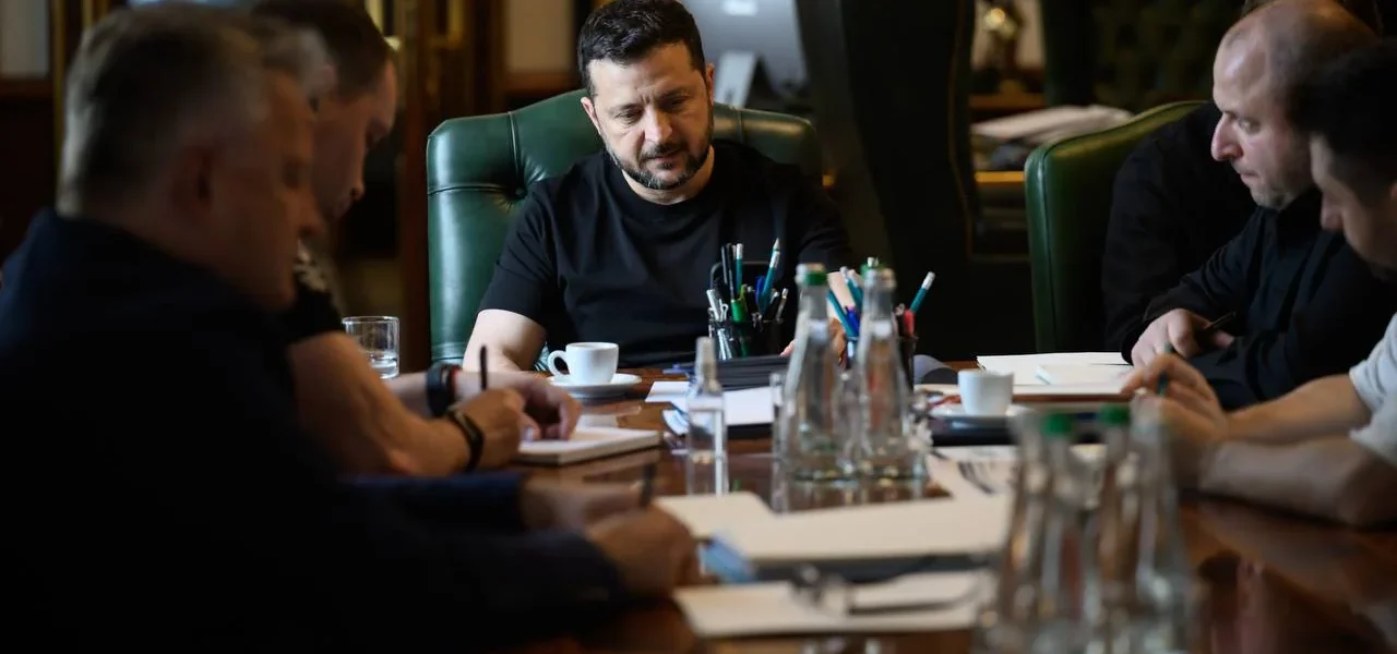 Зеленський у чорній футболці сидить за столом наради в кабінеті з прапором